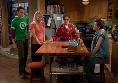 Quatro amigos em uma cozinha. Um homem de camiseta geek, uma mulher loira, um homem de roupão e um de colete jeans interagem casualmente.