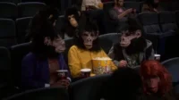Várias pessoas usando máscaras realistas de macaco e cabelo falso assistem a um filme em um cinema escuro, comendo pipoca. Clima divertido.