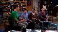 Quatro homens jogam videogame em uma sala de estar cheia de livros e um telescópio. O clima é descontraído e focado.