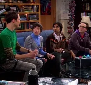 Quatro homens jogam videogame em uma sala de estar cheia de livros e um telescópio. O clima é descontraído e focado.