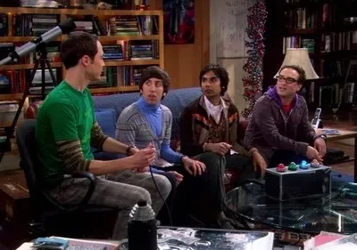 Quatro homens jogam videogame em uma sala de estar cheia de livros e um telescópio. O clima é descontraído e focado.