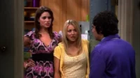 Duas mulheres com expressões de surpresa/choque encaram um homem de casaco roxo, visto de costas, em uma sala.