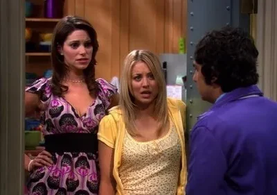 Duas mulheres com expressões de surpresa/choque encaram um homem de casaco roxo, visto de costas, em uma sala.