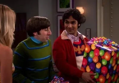 Um homem sorridente oferece um presente colorido de balões. Outro homem de verde e uma mulher loira conversam.