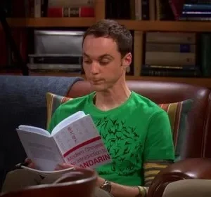 Um homem de camisa verde e cabelo curto lê um livro sobre mandarim em uma poltrona. Uma estante de livros ao fundo.