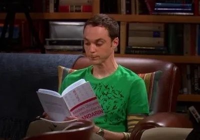 Um homem de camisa verde e cabelo curto lê um livro sobre mandarim em uma poltrona. Uma estante de livros ao fundo.