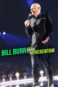 Comediante careca sorridente, camisa escura, em palco iluminado com microfone, gesticulando. Clima de stand-up.