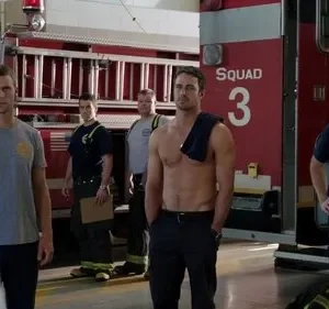 Vários bombeiros sérios em um quartel, com caminhões vermelhos. Um homem está sem camisa, outros com uniforme.