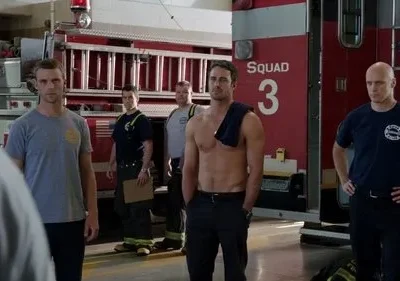 Vários bombeiros sérios em um quartel, com caminhões vermelhos. Um homem está sem camisa, outros com uniforme.