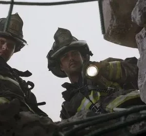 Dois bombeiros equipados olham para baixo com lanternas, em meio a escombros e concreto. Clima tenso de resgate.