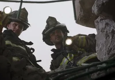 Dois bombeiros equipados olham para baixo com lanternas, em meio a escombros e concreto. Clima tenso de resgate.