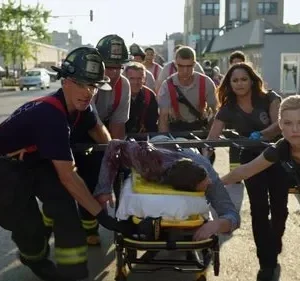 Bombeiros e paramédicos correm por rua urbana, transportando maca com vítima ensanguentada. Clima de emergência e ação.