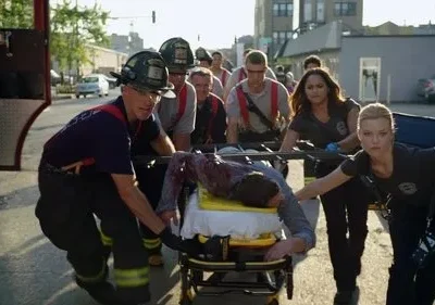 Bombeiros e paramédicos correm por rua urbana, transportando maca com vítima ensanguentada. Clima de emergência e ação.