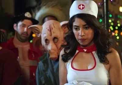 Enfermeira de roupa branca e vermelha com expressão séria em festa. Ao fundo, máscara de porco ensanguentada e outros convidados fantasiados.