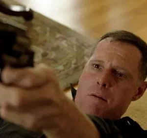 Um homem de rosto sério aponta uma arma escura em primeiro plano. O fundo interior é claro. Clima de ação e suspense.