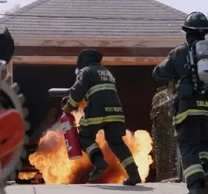 Bombeiros correm com equipamento para combater grandes chamas que saem de uma garagem. Cena de ação e perigo.