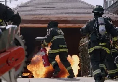 Bombeiros correm com equipamento para combater grandes chamas que saem de uma garagem. Cena de ação e perigo.