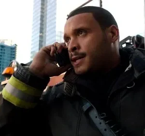 Homem em jaqueta de bombeiro com listras, fala em rádio com olhar sério. Cenário urbano com prédios altos ao fundo.
