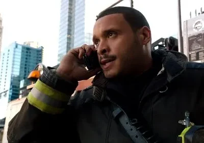 Homem em jaqueta de bombeiro com listras, fala em rádio com olhar sério. Cenário urbano com prédios altos ao fundo.