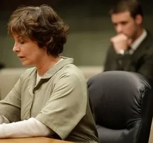 Mulher em uniforme prisional, cabeça baixa e mãos entrelaçadas, em tribunal. Homem de terno pensativo desfocado ao fundo. Clima sério.