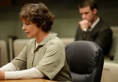 Mulher em uniforme prisional, cabeça baixa e mãos entrelaçadas, em tribunal. Homem de terno pensativo desfocado ao fundo. Clima sério.