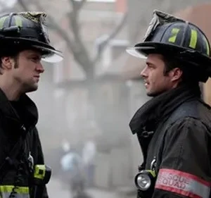 Dois bombeiros com capacetes, vestindo equipamento escuro, se olham intensamente. Fumaça ou neblina ao fundo cria um clima sério.