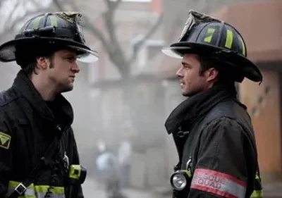 Dois bombeiros com capacetes, vestindo equipamento escuro, se olham intensamente. Fumaça ou neblina ao fundo cria um clima sério.
