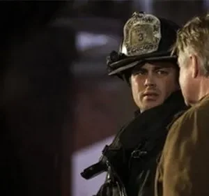 Um bombeiro com capacete olha intensamente para um homem de costas. Cena escura com clima sério e urgente.