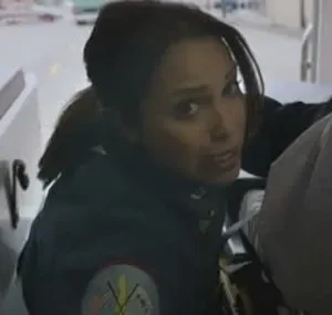 Paramédica com olhar sério em ambulância. Um paciente está na maca ao lado, e o fundo mostra uma rua movimentada.