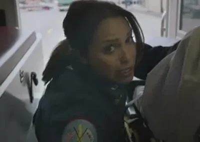 Paramédica com olhar sério em ambulância. Um paciente está na maca ao lado, e o fundo mostra uma rua movimentada.