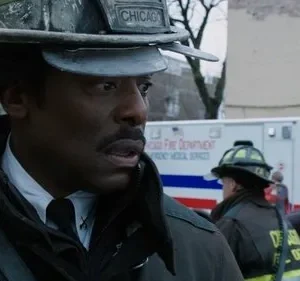 Um bombeiro negro, sério, com capacete Chicago e rádio. Ao fundo, uma ambulância e outros socorristas atendem civis. Cena de emergência.