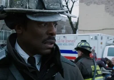 Um bombeiro negro, sério, com capacete Chicago e rádio. Ao fundo, uma ambulância e outros socorristas atendem civis. Cena de emergência.