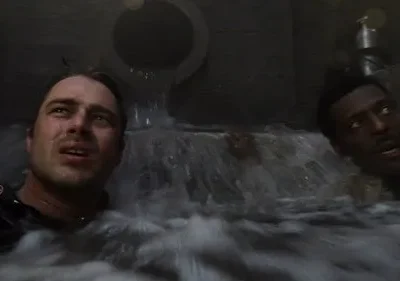 Dois homens assustados em um túnel escuro, submergidos em água turbulenta. Um cano enorme jorra água sobre eles.