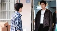Dois homens jovens. Um de camisa xadrez, de costas, perto de caixas. O outro, de jaqueta escura, tem olhar sério em porta 106.