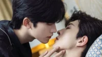 Dois homens jovens em close-up. Um se inclina para beijar o outro, de olhos fechados, que o toca suavemente no rosto. Cena íntima e romântica.