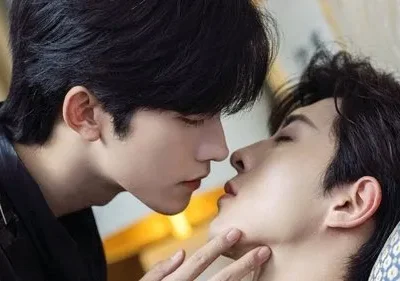 Dois homens jovens em close-up. Um se inclina para beijar o outro, de olhos fechados, que o toca suavemente no rosto. Cena íntima e romântica.