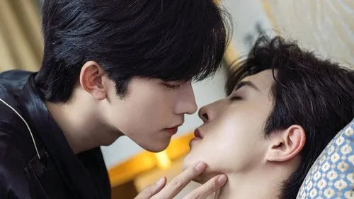 Dois homens jovens em close-up. Um se inclina para beijar o outro, de olhos fechados, que o toca suavemente no rosto. Cena íntima e romântica.