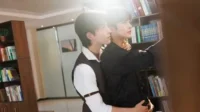 Dois jovens se abraçam em uma biblioteca iluminada. Um estende a mão para um livro. A cena tem um clima romântico e sereno.