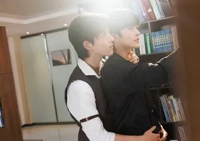 Dois jovens se abraçam em uma biblioteca iluminada. Um estende a mão para um livro. A cena tem um clima romântico e sereno.