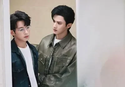 Dois homens jovens se entreolham com expressão séria em um ambiente claro. Um de óculos e jaqueta jeans, o outro de jaqueta verde.