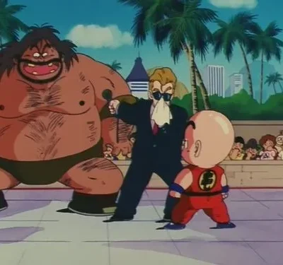 Um gigante peludo sorri diante de um menino careca de uniforme de luta. Um homem de terno com microfone ao lado. Público assiste. Cenário urbano ensolarado.