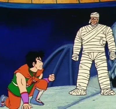 Homem agachado em traje de luta laranja e verde encara um oponente tipo múmia em pé. Fundo escuro com cabeça de dragão de pedra.