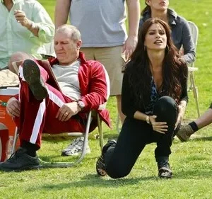 Um homem de agasalho vermelho senta entediado enquanto uma mulher ajoelhada olha intensamente para cima. Cenário externo de gramado com outras pessoas.