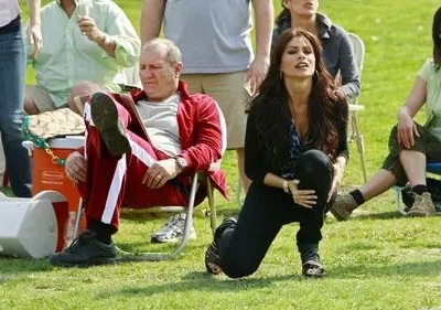 Um homem de agasalho vermelho senta entediado enquanto uma mulher ajoelhada olha intensamente para cima. Cenário externo de gramado com outras pessoas.