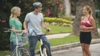 Duas pessoas de bicicleta e uma mulher de roupa esportiva sorriem e conversam em uma rua residencial. Dia ensolarado e alegre.