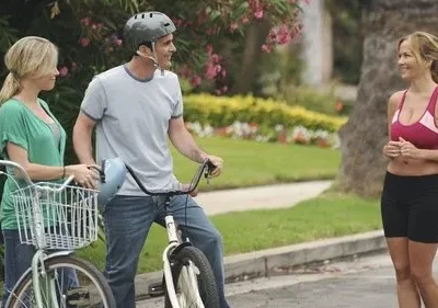 Duas pessoas de bicicleta e uma mulher de roupa esportiva sorriem e conversam em uma rua residencial. Dia ensolarado e alegre.