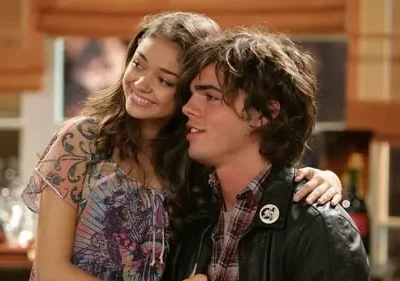 Jovem sorridente com blusa estampada abraça um rapaz de cabelo comprido e jaqueta de couro. Fundo de casa, clima caloroso.