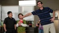 Um homem sorridente tapa a boca de um menino que segura um futebol, enquanto outro menino observa. Humor familiar em casa.