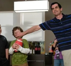 Um homem sorridente tapa a boca de um menino que segura um futebol, enquanto outro menino observa. Humor familiar em casa.
