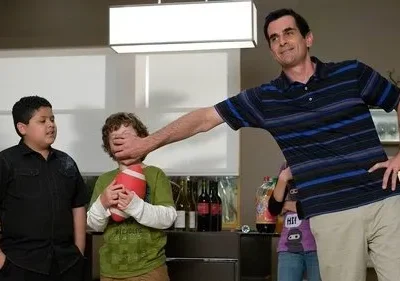 Um homem sorridente tapa a boca de um menino que segura um futebol, enquanto outro menino observa. Humor familiar em casa.